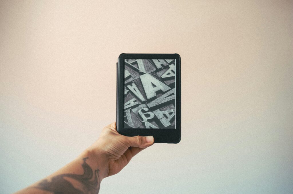 Potetovaná ruka držiaca čítačku Kindle s typografickým screensaverom na E-Ink displeji